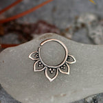 A 925 silver body jewelry piece with intricate detailing sits on a frosted surface. The MAWAR Lotus Flower Septum Ring in Silver by Kaya Movement, an 18-gauge septum nose daith rook helix ring, is adorned with small, petal-like shapes featuring dot patterns inside each petal that give it a floral appearance. The background includes slightly blurred natural elements.