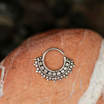 A close-up image of the GAE Mandala Septum Ring in Silver by Kaya Movement showcases its intricate, ornate detailing, including small round beads. The simple tribal ethnic design of this 18-gauge 925 silver body jewelry ring is accentuated as it rests on a reddish-orange stone with white streaks, providing a rustic and earthy background.