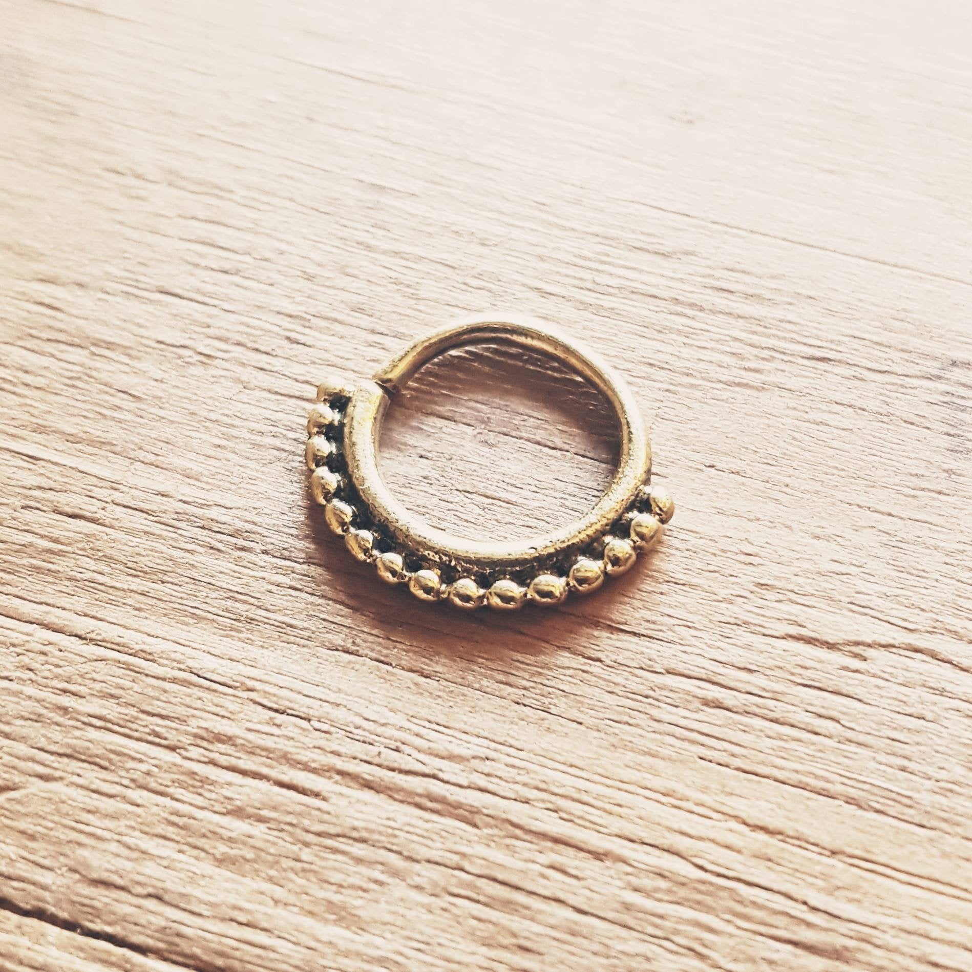The IRI Seamless Septum Ring in Gold by Kaya Movement, featuring a delicate dotted pattern around the lower half, rests gracefully on a wooden surface. The intricate design lends it a tribal ethnic charm, making it an exquisite piece of body jewelry suitable as either a septum nose ring or tiny hoop earring.