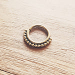 The IRI Seamless Septum Ring in Gold by Kaya Movement, featuring a delicate dotted pattern around the lower half, rests gracefully on a wooden surface. The intricate design lends it a tribal ethnic charm, making it an exquisite piece of body jewelry suitable as either a septum nose ring or tiny hoop earring.