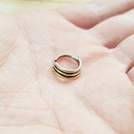 A hand holds the TRIA Septum Ring in Gold by Kaya Movement, an 18 gauge septum ring with a sleek, multi-banded design. The polished brass body jewelry features a seamless closure, displayed against the palm's intricate texture.