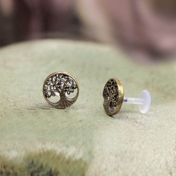 Introducing Tree of Life Labret Studs in Silver by Kaya Movement. These elegant 16-gauge pieces feature an intricate tribal Tree of Life design with sprawling branches. Highlighting the detailed artwork, one labret stud stands upright while the other lies flat, showcasing the clear bio-plastic stopper on the back. Crafted from 925 Sterling Silver, the slightly textured backdrop is purposely blurred for artistic effect.