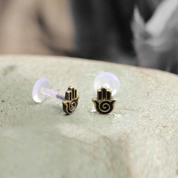 Close-up of a pair of small Kaya Movement earrings, each featuring a minimalist hamsa hand design with a spiral in the palm. The HAMSA HAND Labret Studs, offered in 18 or 16 gauge, are crafted from gold brass and secured with bio-plastic push backs. The earrings rest on a light-colored, textured surface.