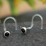 A close-up image showcasing the elegant pair of Kaya Movement's EDEN Nose Screw Studs, available in gold or silver and featuring small, round onyx stones. Each 20-gauge stud is complemented by a curved clear plastic backing that ensures both comfort and a secure fit. The blurred background with hints of green and yellow enhances the sophistication of this exquisite body jewelry.