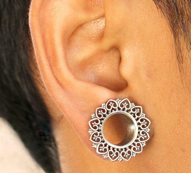 Close-up image of an ear adorned with a circular, intricate filigree earring. This exquisite silver tunnel features an ornate heart and scrollwork design forming a flower-like pattern around a central, open tunnel gauge, part of the stunning ONAS Sacred Lotus Mandala Round Tunnels collection from Kaya Movement.