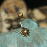 A close-up image showcases the Kaya Movement's exquisite Lotus Mandala Belly Piercing Ring in Gold, a 14-gauge navel jewelry piece crafted from ornate 316L surgical steel with a flower-like design. In the slightly blurred background, skull decorations can be seen, while the elegantly packaged jewelry is beautifully presented on a textured surface.