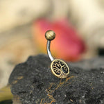 The Tree of Life Belly Piercing Ring in Gold by Kaya Movement is a beautifully curved navel jewelry piece featuring a golden top ball and an intricately detailed tree of life charm at the base. The charm rests gracefully on a dark, textured surface, with a blurred background showcasing earthy tones and hints of red-orange—perfect for elegant gift packaging. This exquisite piece is designed in 14 gauge for timeless sophistication.