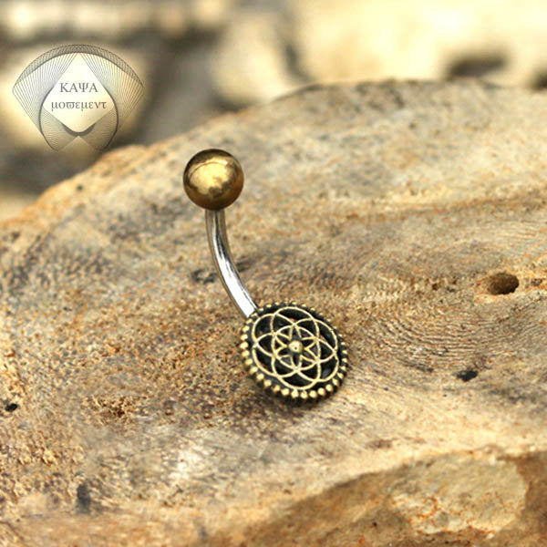 A "Flower of Life Belly Piercing Ring in Gold" from Kaya Movement, featuring a curved surgical steel barbell with a small gold ball on one end and a decorative circular floral pattern on the other end, is placed on a textured rock surface. The background showcases a subtle "KAJA" logo.