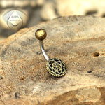 A "Flower of Life Belly Piercing Ring in Gold" from Kaya Movement, featuring a curved surgical steel barbell with a small gold ball on one end and a decorative circular floral pattern on the other end, is placed on a textured rock surface. The background showcases a subtle "KAJA" logo.