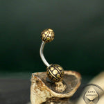 Close-up of a GEO Globe Belly Piercing Ring in Gold from Kaya Movement, featuring two spherical ends. The spheres are textured with gold-colored circular patterns on a dark background. The 14-gauge surgical steel barbell is displayed on a small piece of natural wood. The logo "Kaya Movement" is shown in the bottom right corner.