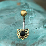 A close-up image of the DORE Butterfly Belly Bar in Gold by Kaya Movement, showcasing a gold-colored ball at the top and a heart-shaped design embellished with intricate golden accents and a central black onyx gemstone, set against a translucent background that mimics natural turquoise stone.