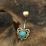 A DORE Butterfly Belly Bar in Gold with Turquoise from Kaya Movement is displayed on a rustic, textured wooden surface. This 14-gauge piece features a rounded turquoise stone encircled by an intricate gold design, connected to a sleek surgical steel ball at the end of its curved bar.