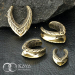 A display of four intricately designed MYRI Tribal Saddle Tunnels in gold, ranging from 12mm to 25mm gauge, is set against a dark, stone background. The tunnels feature detailed ethnic patterns and leaf-like motifs. The "Kaya Movement" logo and name are visible in the bottom left corner.