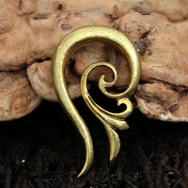 The TILA Spiral Ear Hangers in Gold by Kaya Movement, a 2 gauge ear accessory made from yellow brass, sits against a rustic, textured wooden background. Featuring an intricate, curving design resembling a stylized ear, these ear hangers boast smooth, flowing lines and a polished finish that enhances their feminine aesthetic.