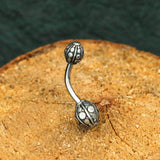 A close-up shot of the Kaya Movement's GEO Globe Belly Piercing Ring in Gold or Silver, featuring two ornate, spherical ends with intricate designs. The 14 gauge navel jewelry is placed on a textured wooden surface against a dark background.