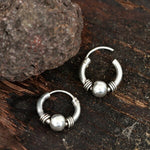 A pair of Kaya Movement's Huggie Hoop Earrings, Silver 925, featuring ornate tribal designs rests on a dark, rustic wooden surface. One ethnic body jewelry earring is fully closed while the other is slightly open. A rough, textured rock is positioned above the earrings on the top left.