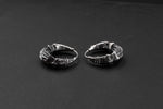 The Kaya Movement's Silver Futuristic Hoop Earrings, designed for everyday wear and inspired by cyberpunk aesthetics, are elegantly displayed on a solid black background. These unisex earrings feature intricate textured designs with small detailed embellishments and slightly asymmetrical patterns, giving them a handcrafted appearance perfect for tech lovers.