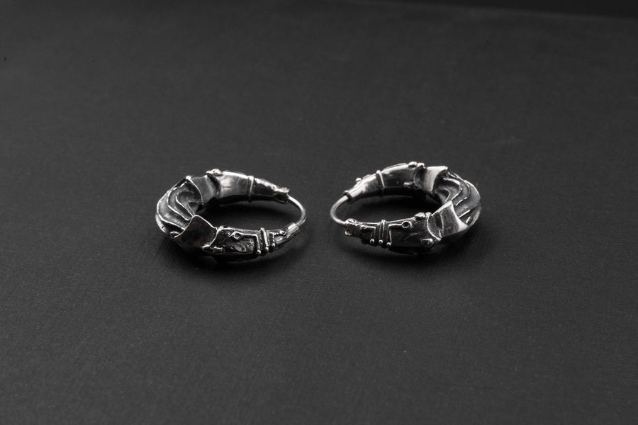 A pair of Bio Organic - Silver Hoop Earrings from Kaya Movement is displayed on a black surface. These intricately designed earrings, featuring detailed patterns and textured elements, exhibit a distinct cyberpunk style inspired by futuristic and Alien Xenomorph aesthetics. They present an artistic statement with small decorative components that add to their unique, Hr Giger-inspired look.