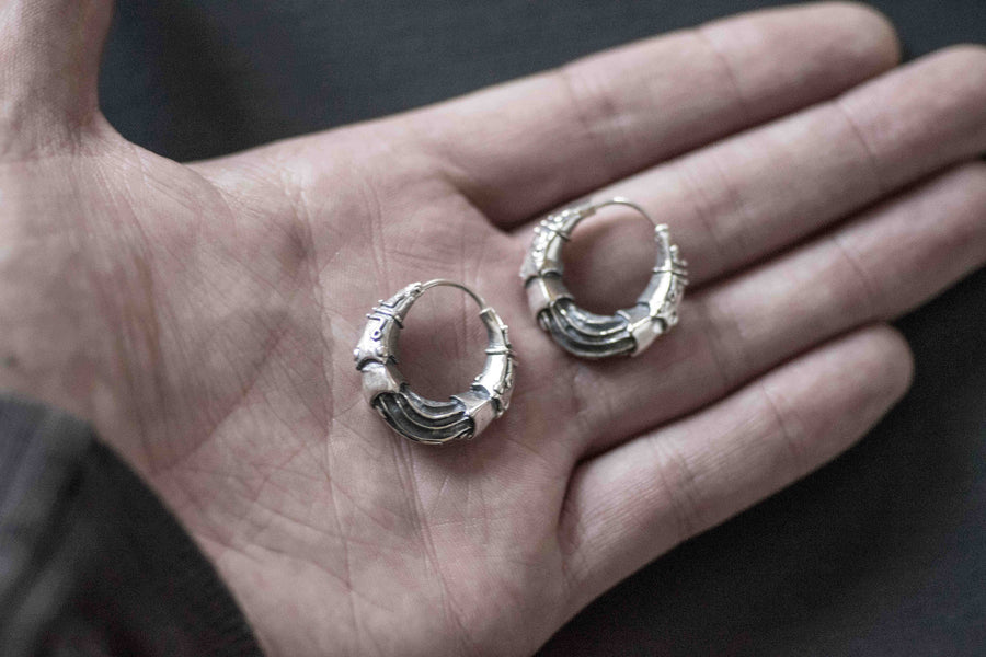A person holds a pair of intricate, futuristic Bio Organic silver hoop earrings by Kaya Movement in their palm. Each earring showcases detailed, textured designs reminiscent of an alien xenomorph or the intricate biomechanical works of H.R. Giger. The background is blurred, focusing attention on the hand and earrings that evoke a striking cyberpunk aesthetic suitable for making a bold statement.