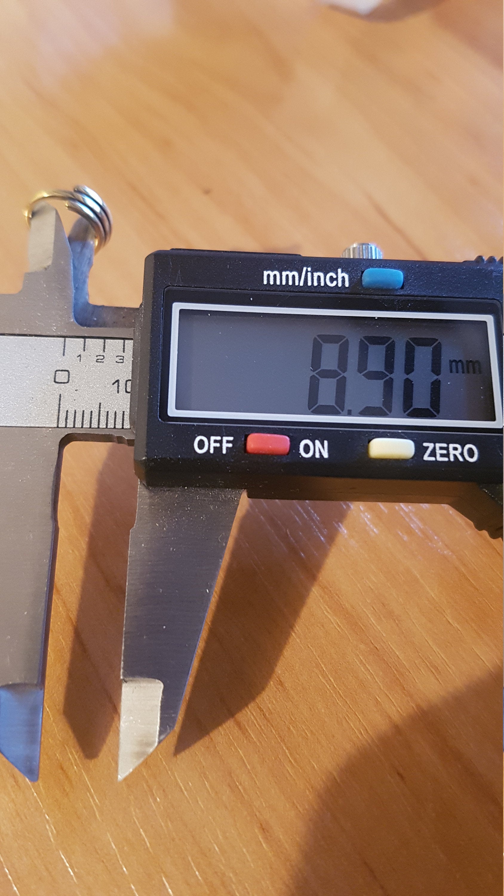 A digital caliper is measuring the width of a small, multi-banded TRIA Septum Ring in Gold by Kaya Movement, which reads 8.90 mm on its electronic display. The measurement is being taken on a wooden surface.