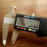 A digital caliper measures a small 18-gauge metal object, displaying a measurement of 9.04 millimeters on its screen. The caliper is held over a wooden surface, with its "on" button illuminated red, perfect for ensuring precision while crafting the Kaya Movement AYM Seamless Septum Ring in Gold for body jewelry.