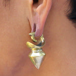 Close-up of a person's ear wearing Kaya Movement's DAYAK Heavy Tribal Ear Weights in Gold, size 0 gauge. The earring features a shiny metallic finish, with an intricate twisted design at the top and a geometric, cone-like shape hanging below. The person's short hair and skin are visible in the background.
