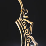Close-up image of an intricately designed, gold-colored Lotus flower mandala earring from Kaya Movement, featuring swirls and loops against a dark background. The handmade brass jewelry is slightly out of focus, emphasizing its delicate curves and craftsmanship.