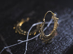 A close-up of a Kaya Movement Lotus Flower Pair Earring, a piece of handmade mandala brass jewelry, lying on a dark, textured surface. The earring boasts intricate patterns and detailing, illuminated by soft lighting that highlights its craftsmanship, with part of another similar earring visible in the blurred background.