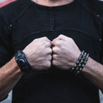 Close-up of a person's clenched fists held together in front of them. The person is wearing a dark shirt and two wristbands: one with a large dark stone and the other with metallic studs, crafted using the lost wax process. Completing the ensemble is an adjustable unisex KRAKO Reptilian Spine Cuff Bangle in Black, from Kaya Movement. The background is blurred.