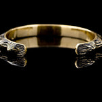 The KRAKO Reptilian Spine Cuff Bangle by Kaya Movement is featured against a black backdrop. This unisex piece, available in gold, showcases textured and intricate designs on both ends. The polished and reflective surface highlights its simple yet elegant aesthetic. Meticulously crafted using the lost wax process, this bracelet offers an adjustable size for a perfect fit.