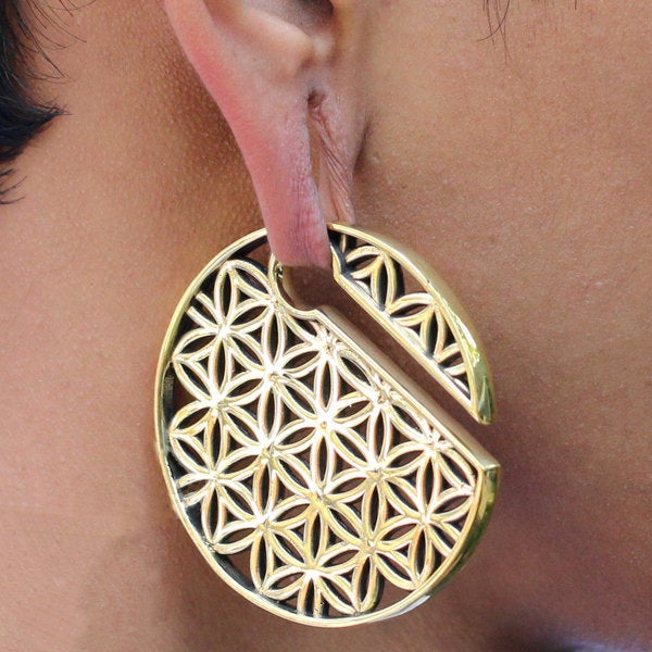 A close-up of a person's ear adorned with Kaya Movement's FLOWER OF LIFE Disc Ear Weights in Gold, designed for 00 gauge. The large, circular gold earring features an intricate geometric pattern that resembles the flower of life and includes a unique design with a small open section. The person's warm-toned skin highlights the earring's exquisite details.