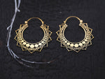 A pair of Lotus Flower earrings from Kaya Movement rests on a dark, textured surface. These handmade brass wire earrings feature intricate mandala designs with circular and crescent-shaped patterns along the edges, giving them an elegant and traditional appearance. A thin white branch is visible to the left.