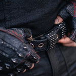 Close-up of a person wearing Kaya Movement's DZALA Black Genuine Leather Fingerless Gloves with red accents and stud knuckles, fastening a black belt adorned with silver studs. The person is dressed in black textured fabric and seems focused on adjusting the belt.