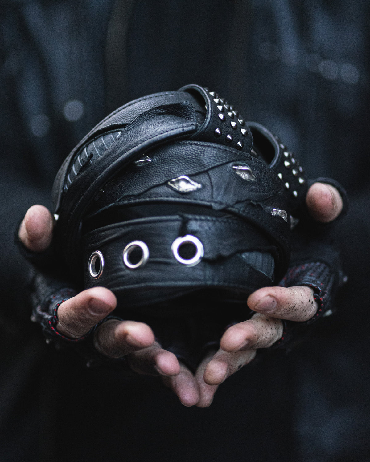 Close-up view of a person wearing fingerless gloves holding the Kaya Movement KRAV Wasteland Men's Black Studded Leather Belt with Silver Spine Studs. The belt appears worn and is held with both hands, set against a dark, blurred background.