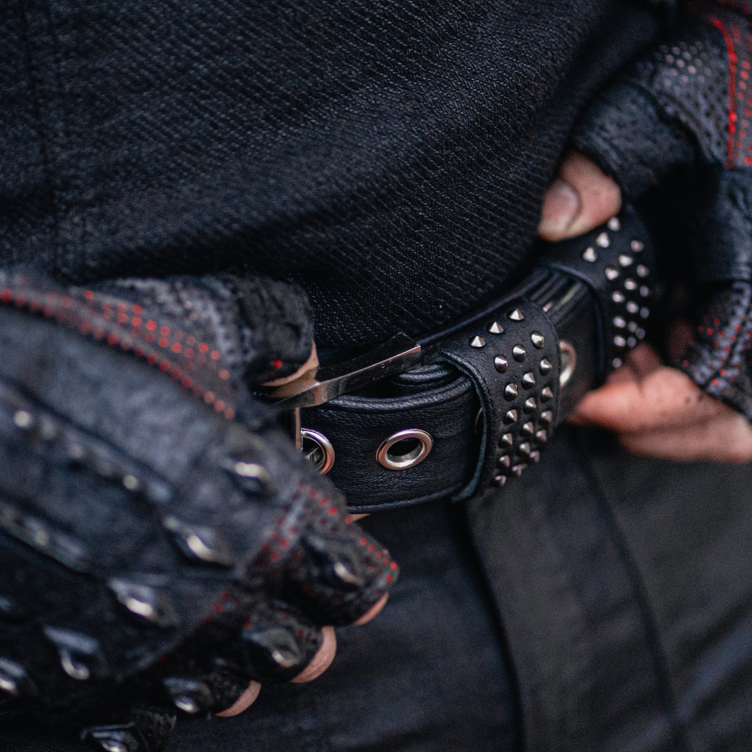 A close-up image captures a person wearing black fingerless gloves with red stitching, adjusting the Kaya Movement KRAV Wasteland Men's Black Studded Leather Belt featuring striking silver spine studs. The attire exudes a rugged and edgy vibe, with the gloved fingers gripping the custom leather goods around their waist.
