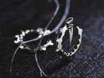 Close-up of two pieces of GIGER Biomechanical Open Ended Hoop Ear Weights in Silver by Kaya Movement on a dark surface. One features a circular design with jagged edges and an inward-extending sharp element. A thin, string-like element wraps around both pieces, adding contrast to the metallic texture, evoking a 0 gauge post-apocalyptic aesthetic.