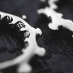 A close-up, monochrome image of intricately designed metallic objects, possibly the GIGER Biomechanical Open Ended Hoop Ear Weights in Silver by Kaya Movement (0 gauge), on a dark surface. The ear weights have curved edges and a textured pattern, suggesting an elegant and artistic style. The focus is on the delicate details of the metalwork.