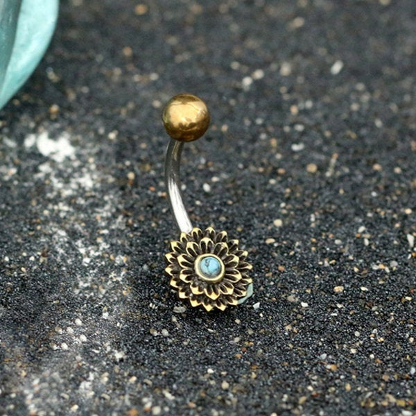 The MADA Lotus Flower Belly Piercing Ring in Gold & Turquoise by Kaya Movement features an exquisite bronze sunflower design with a small turquoise stone center, set against a textured dark gray background. Expertly crafted from surgical steel, the ring has a gentle bend and is adorned with a plain gold top ball. This unique piece of navel jewelry adds elegance to any look.