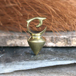 A small metal figurine with a polished, golden finish sits on a stone surface. The object, resembling the DAYAK Heavy Tribal Ear Weights in Gold by Kaya Movement, has a pointed, conical base and a curving, hook-like shape on top that looks like a stylized eye. The background is blurred, showing earthy and textured materials.