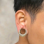 A close-up of a person's ear adorned with Kaya Movement's Reptilian Skin Horseshoe Saddle Tunnels in gold, showcasing a thick, textured, ribbed design reminiscent of reptile skin. The person has short, dark hair against a blurred and neutral-colored background.