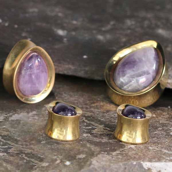 A set of exquisite jewelry featuring Kaya Movement's Amethyst Teardrop Stone Saddle Plugs in Gold, available in gauges from 12mm to 25mm, paired with smaller rounded purple gemstones also set in gold. The pieces are elegantly displayed on a textured dark surface.