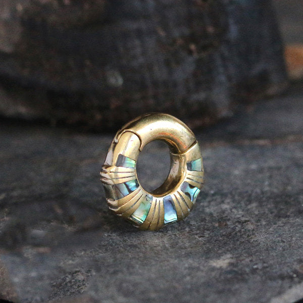 A close-up of Kaya Movement's AVI Clicker Hoop Ear Weights in Gold & Abalone Shell, showcasing their intricate detailing and inlaid iridescent abalone shell segments. The ear weights are set against a dark, textured background, possibly stone or aged wood, highlighting the exquisite contrast between earthy tones and bright colors of these 2 gauge gold-brass beauties.