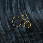 Two ORA Beaded Nose Rings in Gold (16 gauge) and one larger ORA Beaded Nose Ring in Gold (16 gauge) by Kaya Movement are displayed against a dark, textured background. The intricate beadwork adds detail and elegance to these beautiful nose rings inspired by tribal ethnic design.