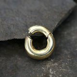 A close-up image of the DAYA Minimalist Hoop Ear Weights in Gold with a clicker by Kaya Movement, featuring a smooth and polished finish. The 2 gauge earring rests on a textured, dark gray stone surface—perfect for those seeking stylish unisex body jewelry.