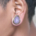 Close-up of a person's ear adorned with the Kaya Movement's Amethyst Teardrop Stone Saddle Plug in Gold. The elegant body jewelry showcases a pale lavender natural amethyst stone set in a gold bezel, perfectly complementing the person's light brown skin tone and short dark hair. This stylish gauge earring is available in sizes from 12mm to 25mm.