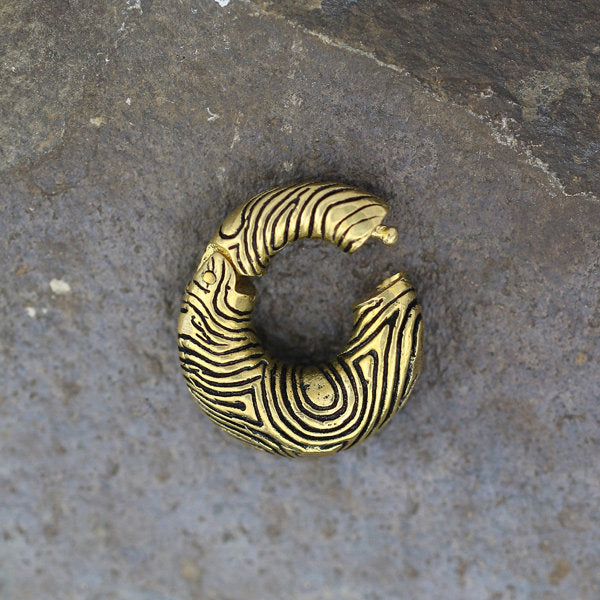 A close-up of the UMI Hoop Ear Weights in Gold with Clicker (2 gauge) by Kaya Movement, showcasing their intricate, organic swirling patterns and incomplete circular shape. The gold ear weights are placed on a textured, grayish stone surface.