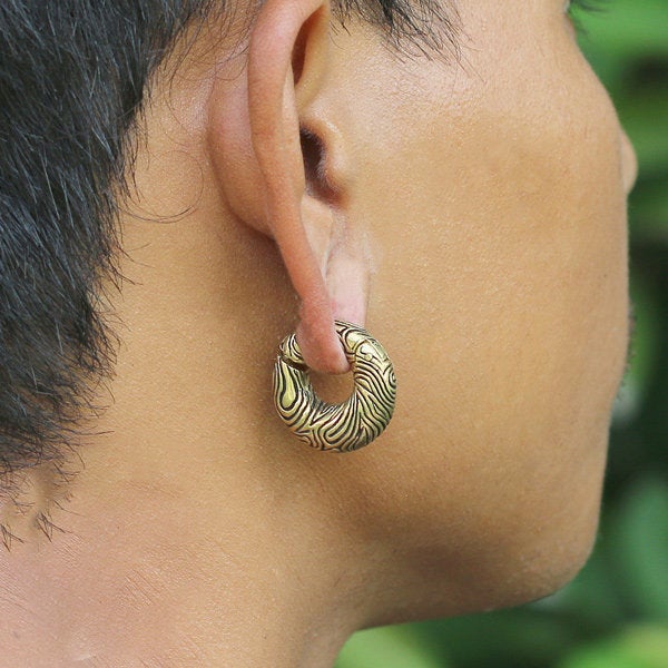 Close-up of a person wearing the UMI Hoop Ear Weights in Gold with Clicker by Kaya Movement, stylishly adorning their stretched earlobe. The elegant design in gold and black resembles wood grain or marble patterns. The organic grooves in these 2 gauge ear weights add an earthy touch, complemented by the wearer’s short hair and a green blurred background visible in profile.