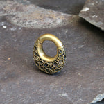 A small, oval-shaped SHAL Hoop Ear Weight in gold with a textured, honeycomb-like pattern from Kaya Movement is positioned on a rough stone surface. The piece, reminiscent of antique brass locks, is closed and boasts an intricate design that enhances its decorative appeal.