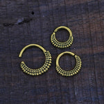 Close-up image of three ISSA Seamless Septum Rings in Gold by Kaya Movement, intricately designed and arranged on a wooden surface. These 16 gauge pieces feature a textured pattern of small, raised bumps, giving them a vintage, handcrafted appearance. Two rings are of identical size while one is slightly larger.