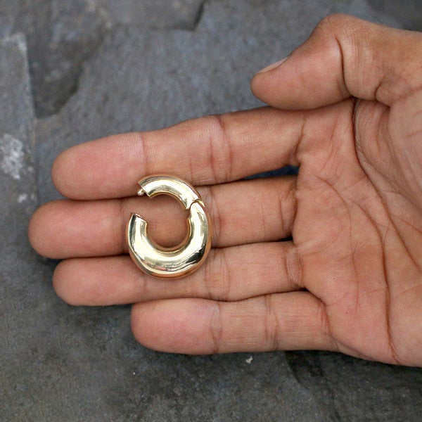 A hand is holding the DAYA Minimalist Hoop Ear Weights in Gold with Clicker by Kaya Movement. The 2 gauge, unisex body jewelry has an oval shape with a smooth, shiny surface. The background is a textured, gray stone surface.